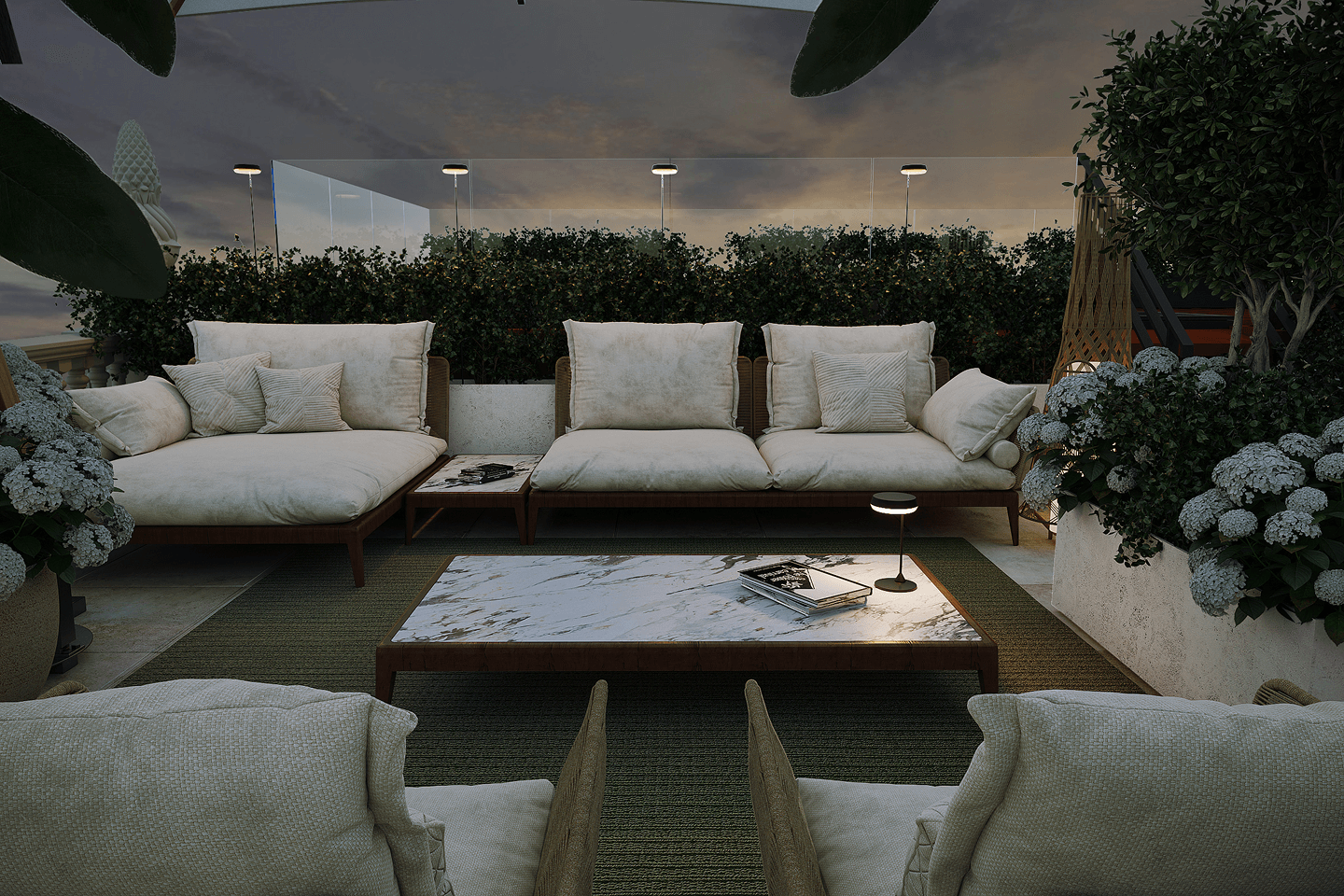 Outdoor seating area with beige cushioned sofas, a marble-top coffee table with books and a small lamp, surrounded by greenery and flowering plants at dusk.